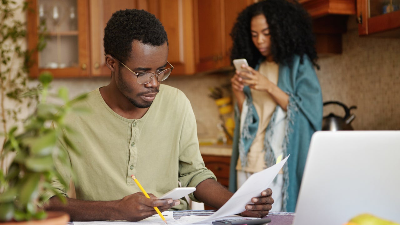 Man sits at kitchen table with pen and paper while woman stand behind on cell phone