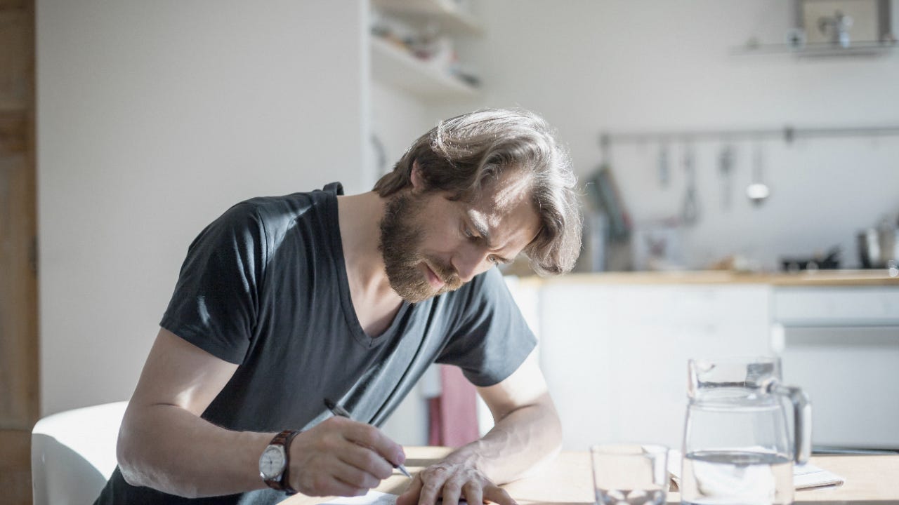 A bearded white man sitting at a table at home filling out a document with a pitcher and glass on the table in front of him.