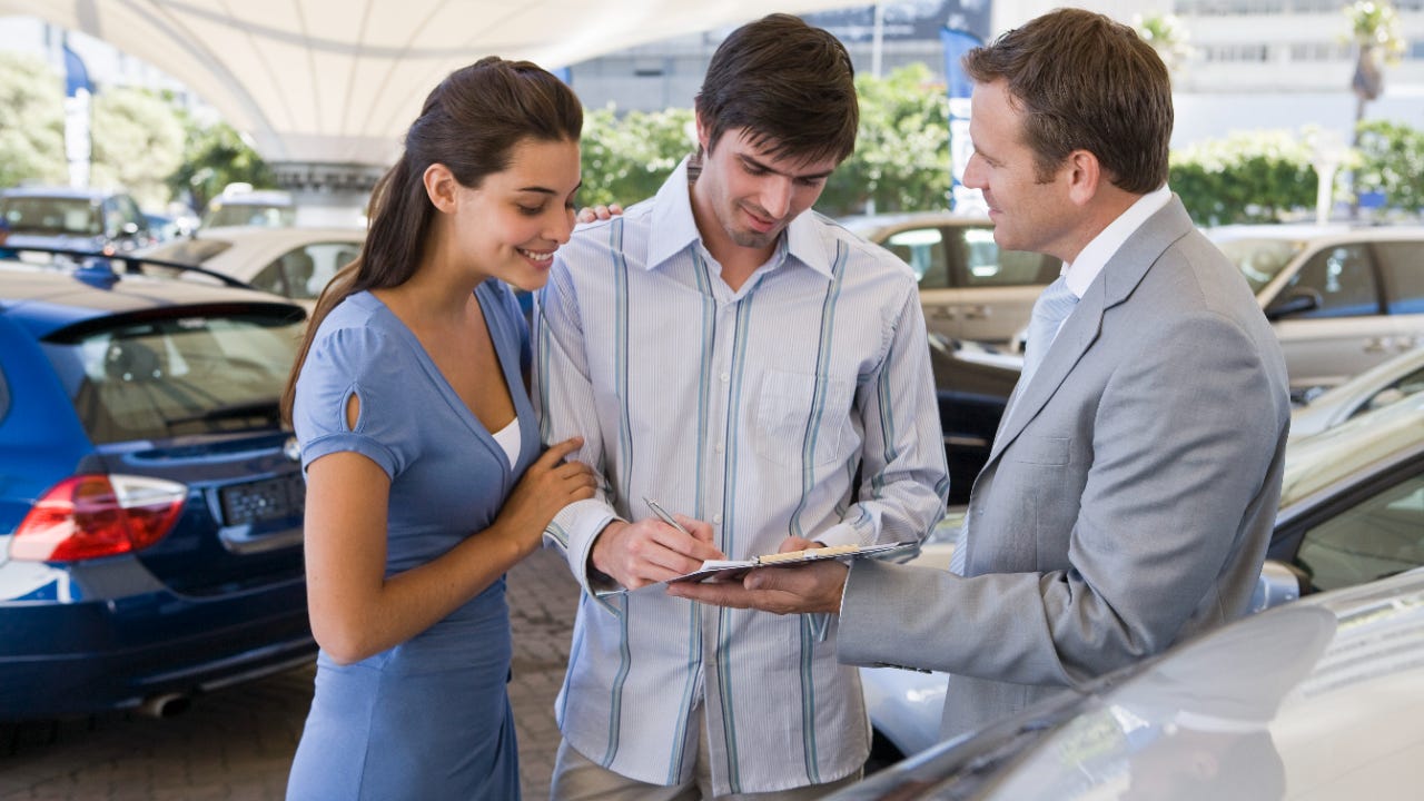 Young white man and woman standing with a car salesperson, signing a document