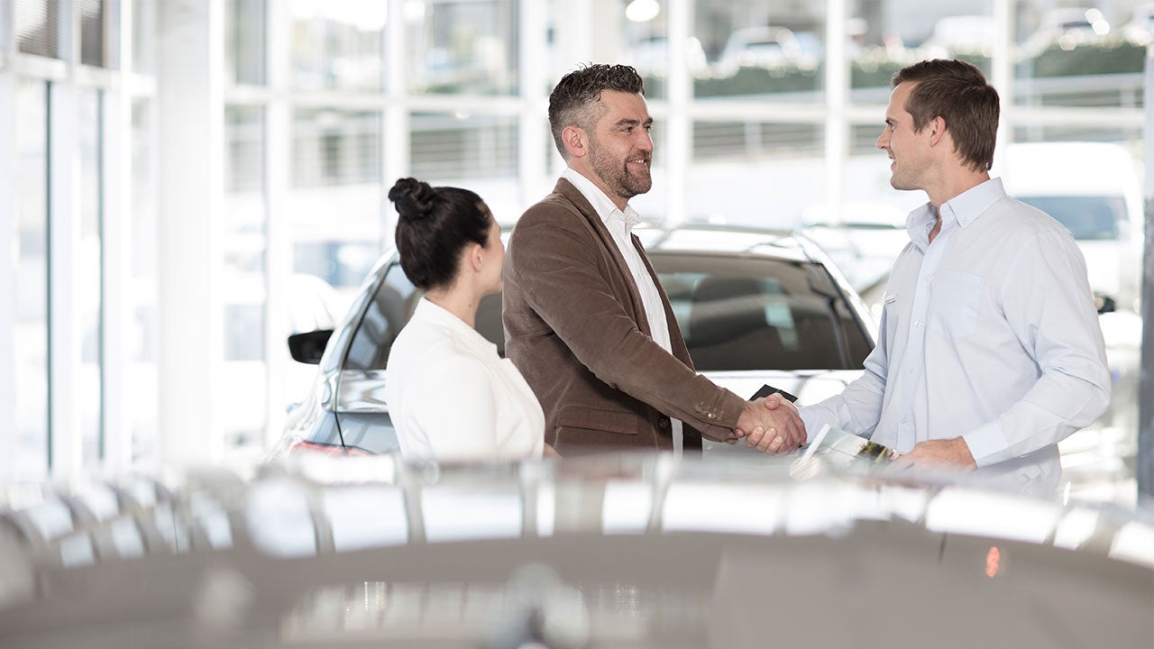 Man and woman at a car dealership talking to a salesperson. The man is shaking hands with the salesperson.