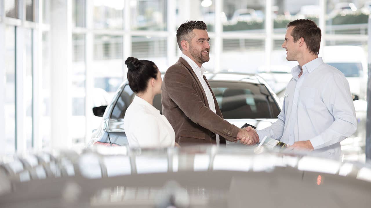 Man and woman at a car dealership talking to a salesperson. The man is shaking hands with the salesperson.