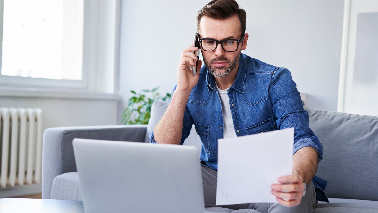 A white man using his phone. He has a piece of paper in his other hand and an open laptop in front of him.