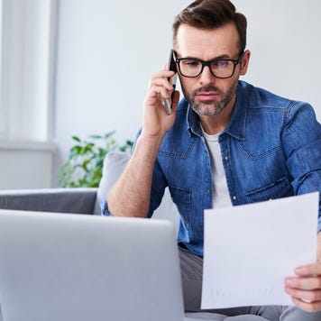 A white man using his phone. He has a piece of paper in his other hand and an open laptop in front of him.
