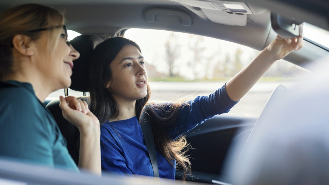 A teenage girl is adjusting the rearview mirror in her car while her mom is sitting in the passenger seat.