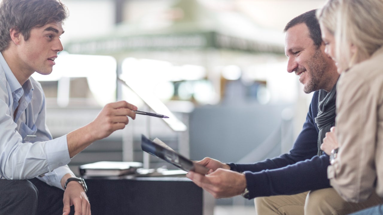 Three people sitting together with a blurred background, one man on the left pointing with a pen and a man and a woman on the right smiling and reviewing a paper