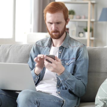 Red haired man sits on white sofa with laptop on knees and scrolls phone, looking pensive