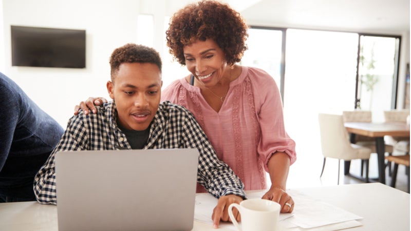 Middle aged woman helps teenage son working on laptop