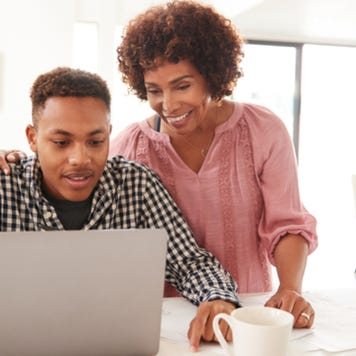 Middle aged woman helps teenage son working on laptop