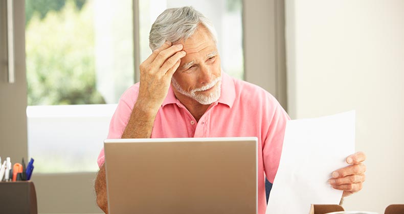 Worried senior man in pink shirt reading paper, laptop on table | iStock.com/monkeybusinessimages