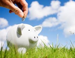 A hand putting a coin in a white piggy bank that is standing on the grass.