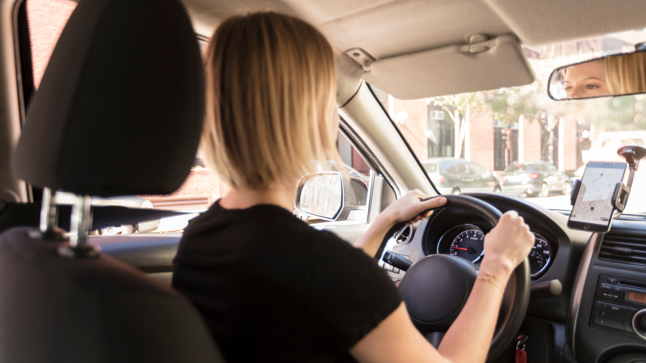 View from passenger in back seat of beautiful young blond woman driver, chauffeur driving a car with directions on her phone on a beautiful sunny day, Commute to work or to airport