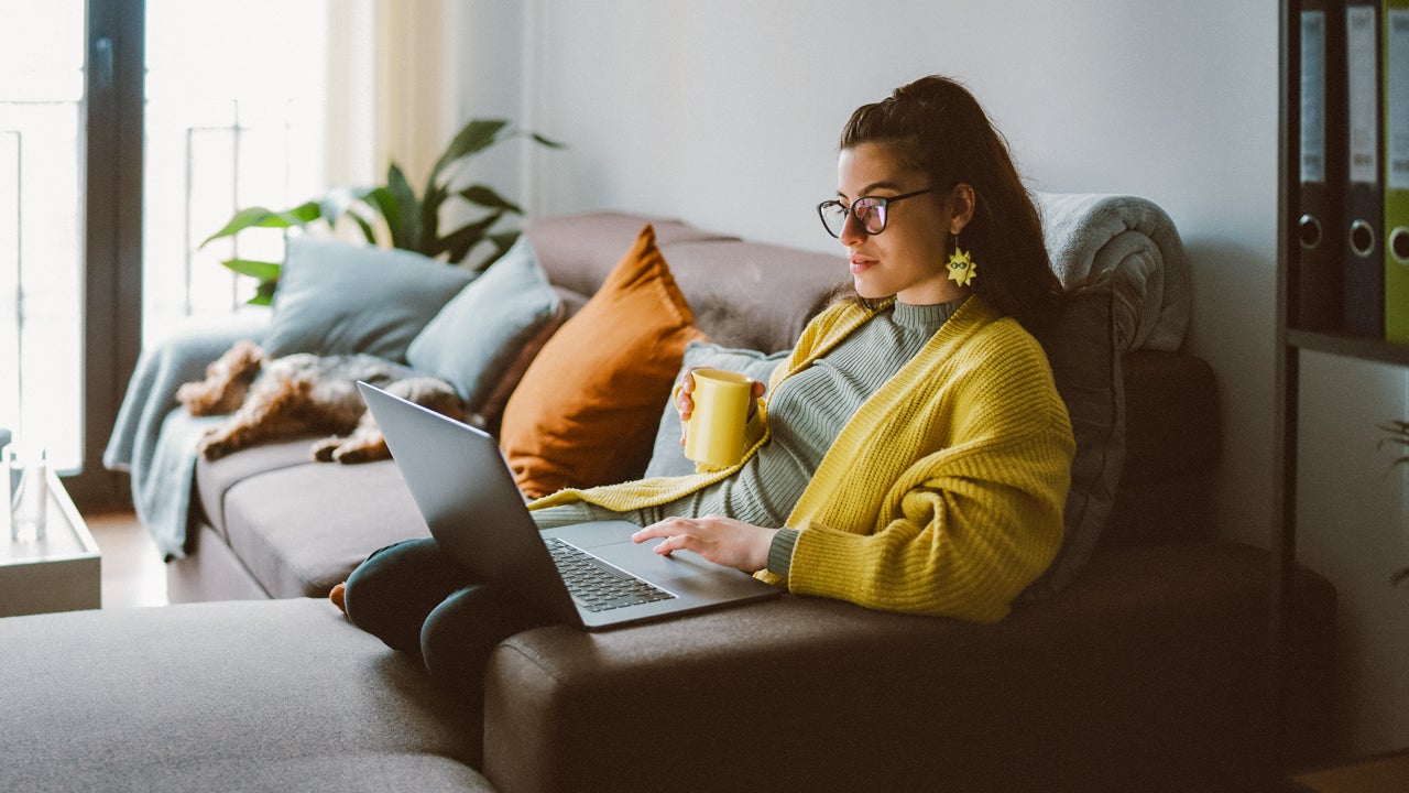 A white woman holds a mug while sitting on the couch and browsing a laptop.