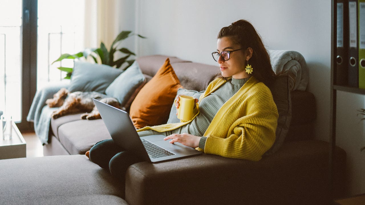 A white woman holds a mug while sitting on the couch and browsing a laptop.