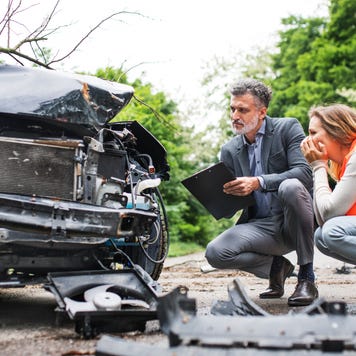A woman crouches, wearing an orange vest and examining the damage to the front of her car while an insurance agent with a clipboard performs an inspection.