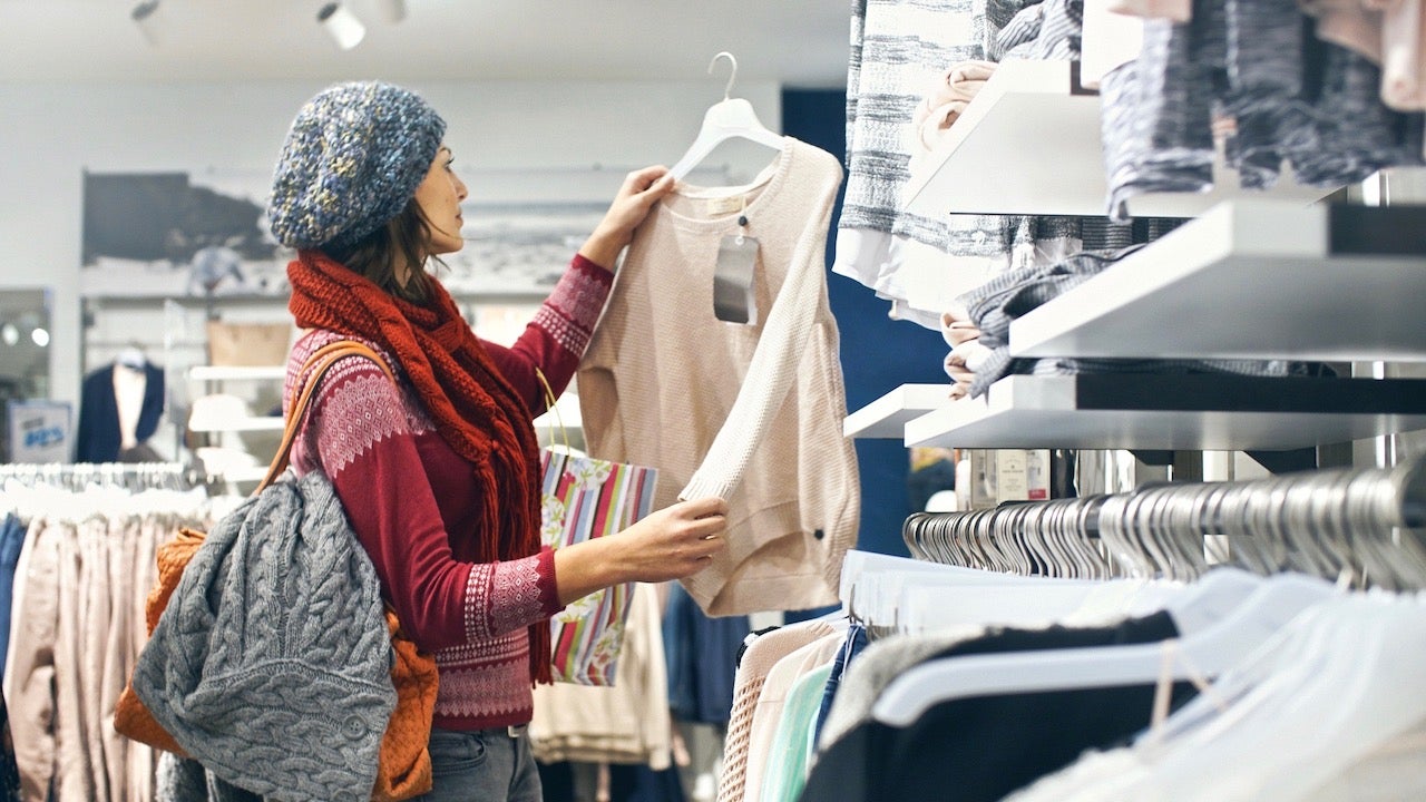 Closeup of smiling blond woman choosing clothes at department store  in local supermarket. She’s holding a beige blouse and looking at it. The woman is wearing gray cap, red sweater and scarf. Side view.