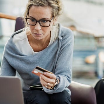 Middle-aged woman with glasses holds credit card while working on laptop with a suitcase nearby