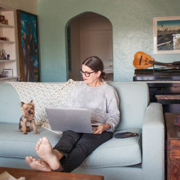 A woman with a laptop sits on a couch with a small dog