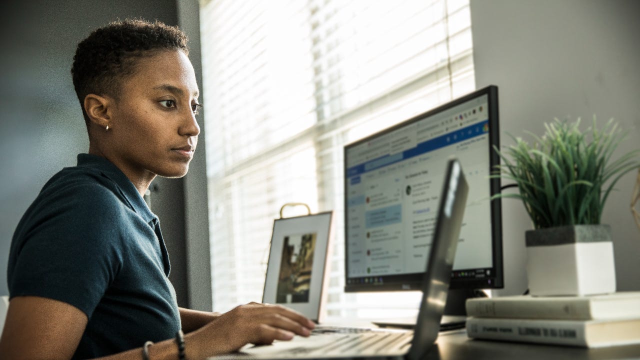 A young woman works on several computers at a home office