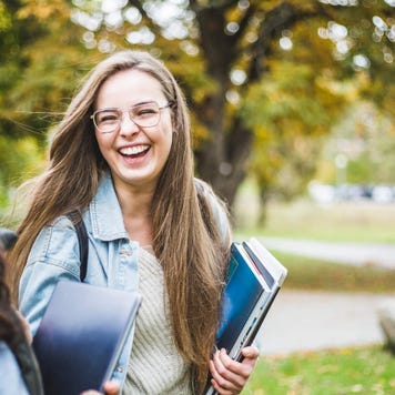 Two happy female students walk and laugh on a university campus with books in hand