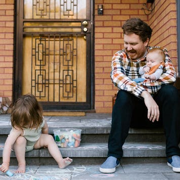 A parent sits on the stoop in front of their home with one baby child in their arms and another, toddler, drawing with chalk on the walkway.