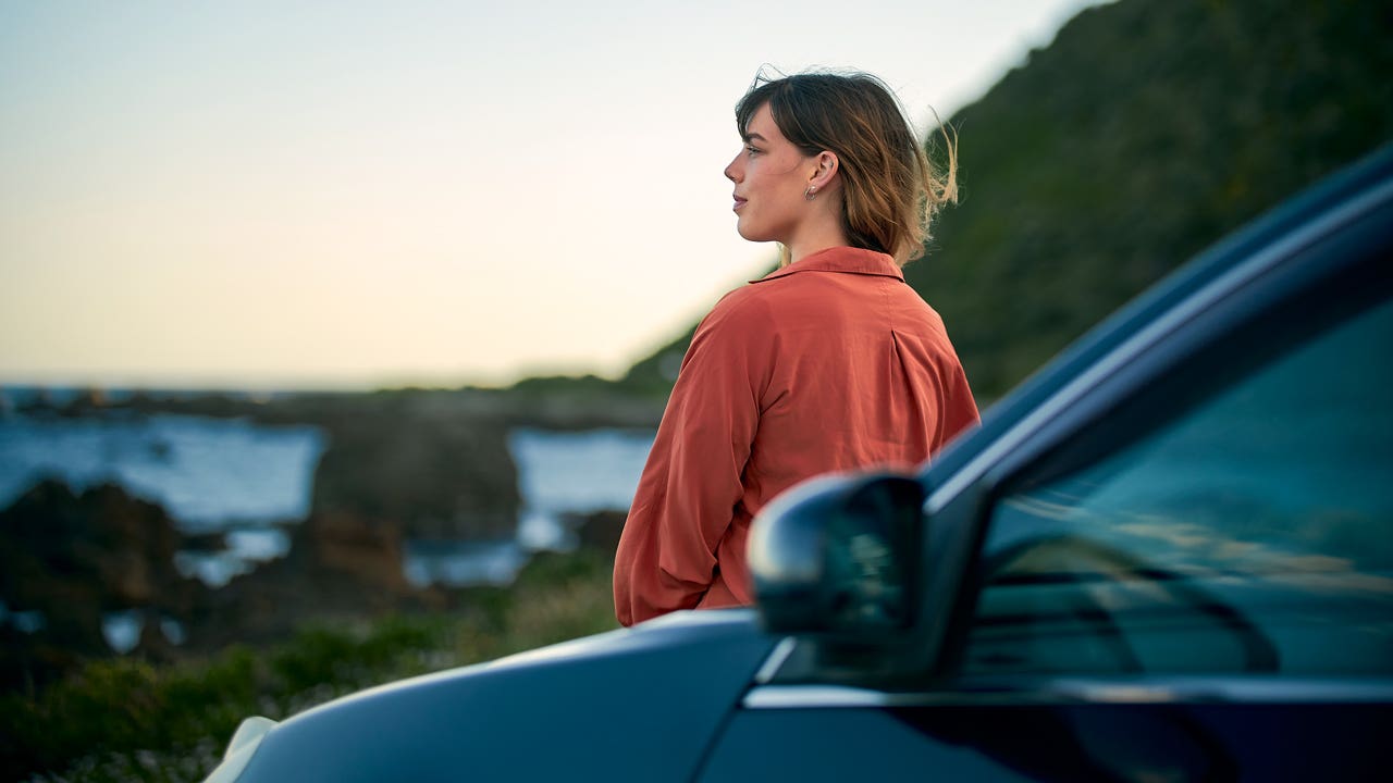 An adult stands by their car and stares out at a nice scene as the sun sets in the distance.