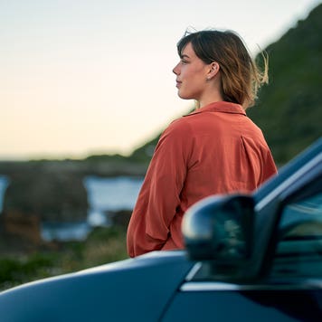 An adult stands by their car and stares out at a nice scene as the sun sets in the distance.