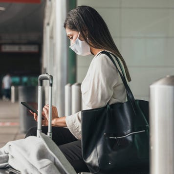 Young woman sitting outside airport terminal and wearing a mask uses her smartphone while surrounded by her luggage