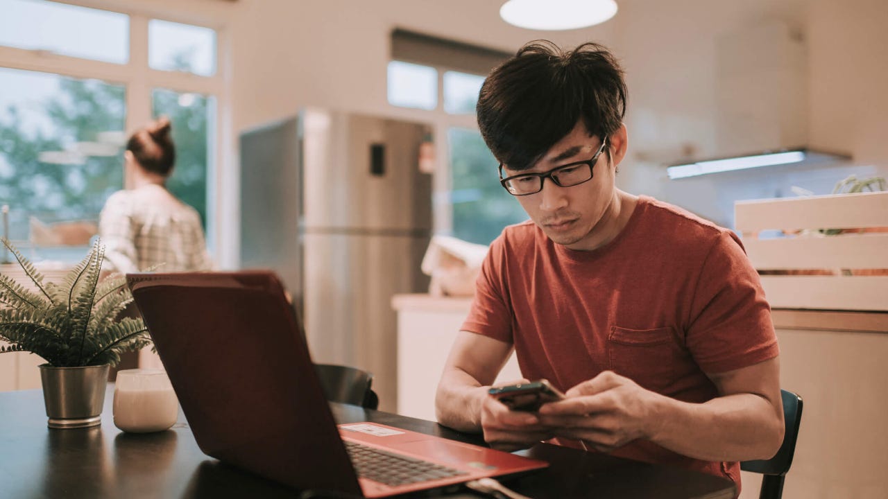 Young man works at kitchen table with laptop and smartphone