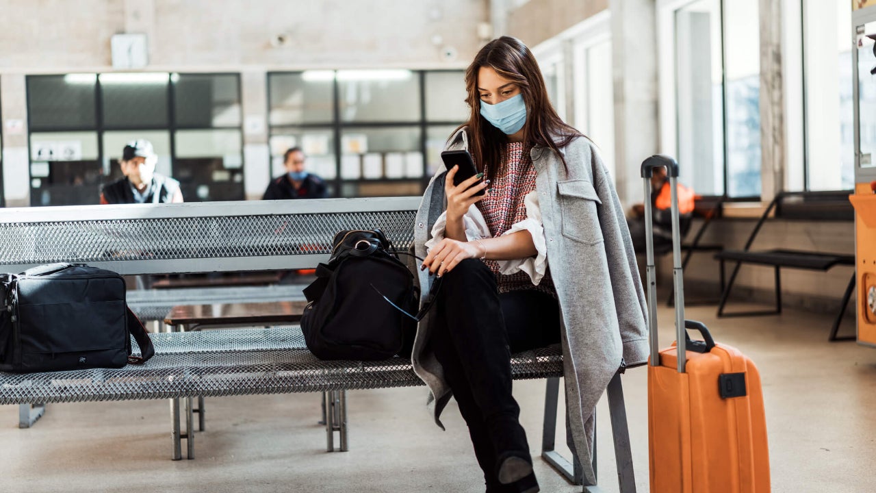 woman witha. mask sitting at an airport terminal with her phone in hand