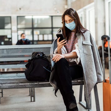 woman witha. mask sitting at an airport terminal with her phone in hand
