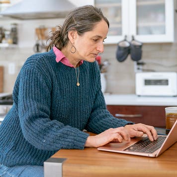 A mid adult woman sits at the counter in her kitchen and uses a laptop computer for online banking while managing her personal finances.