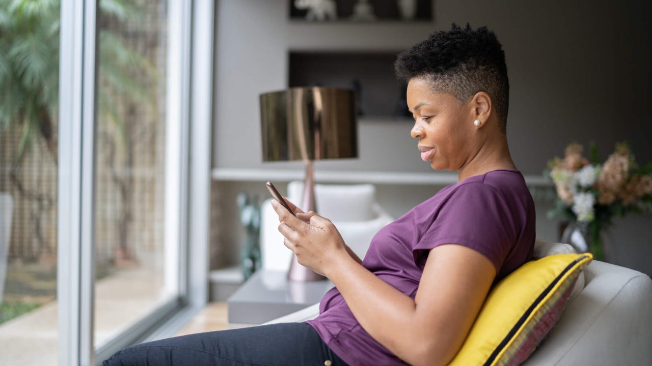 Adult woman sits on the couch in front of her living room window as she uses her smartphone