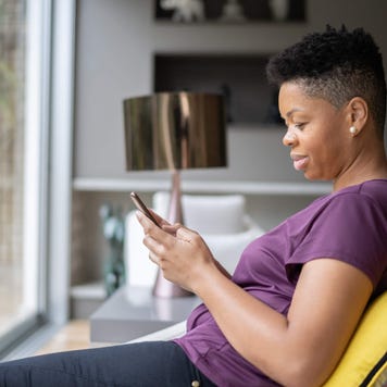 Adult woman sits on the couch in front of her living room window as she uses her smartphone