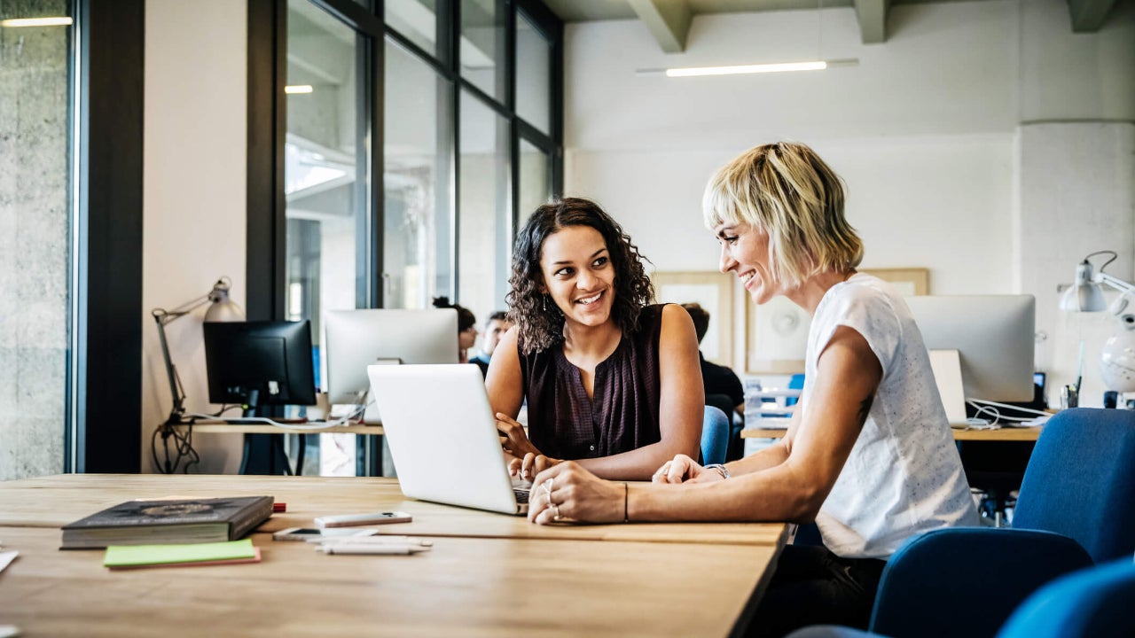 Two casual businesswomen sit at a conference table and view a laptop screen together while smiling