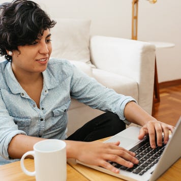 Woman sitting on floor and working on laptop. Female sitting at table with cup of coffee and using laptop.