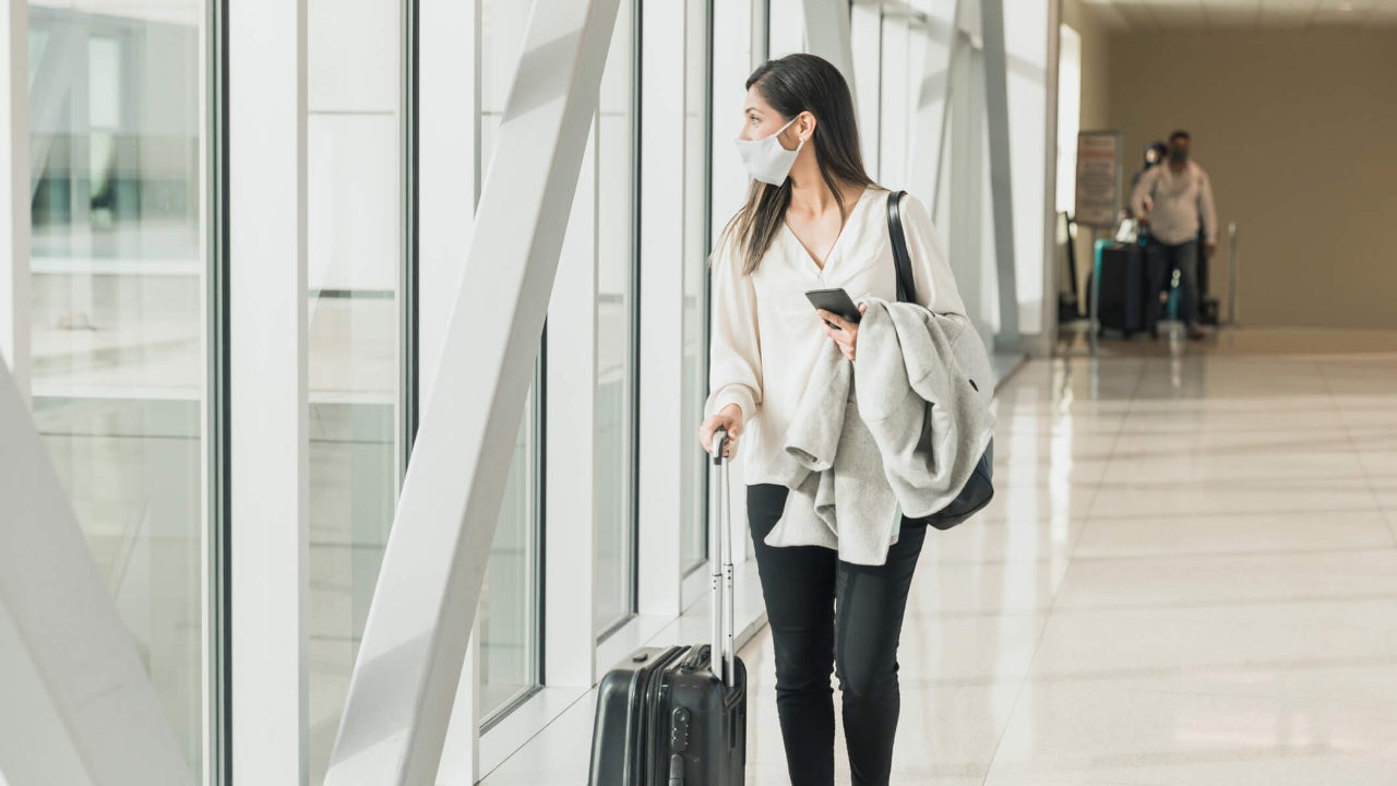 Young adult woman walks down concourse with suitcase, mask and phone