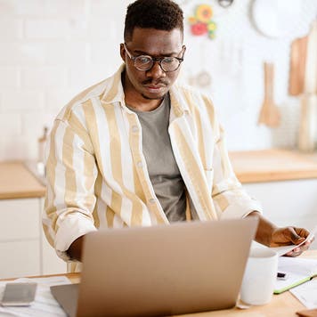 African man working from home, using laptop, paying utility bills. Young man doing taxes at home. Man talking to costumer service.