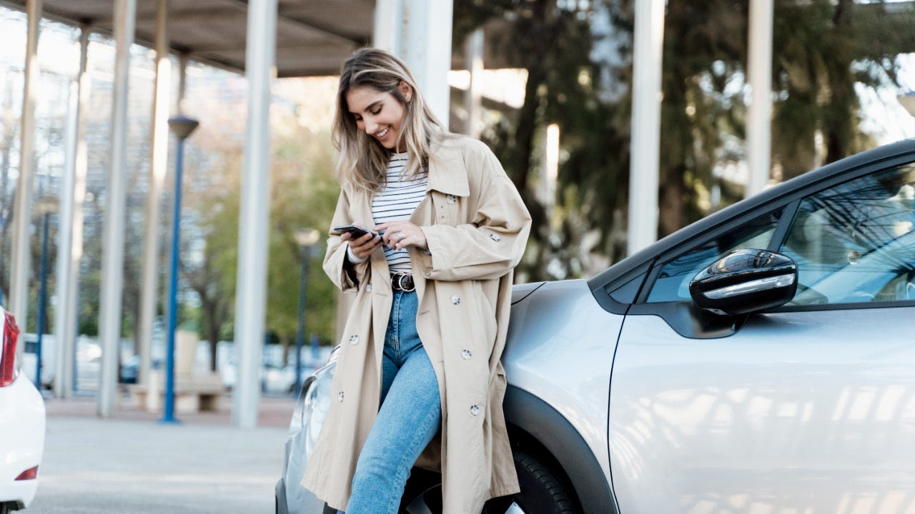 Smiling woman in beige overcoat looking at phone while leaned against silver car