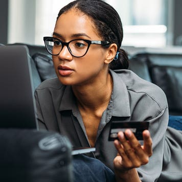 woman laying on couch while working on laptop and holding credit card