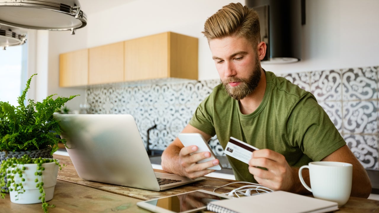 man sitting in kitchen looking at his phone and holding credit card