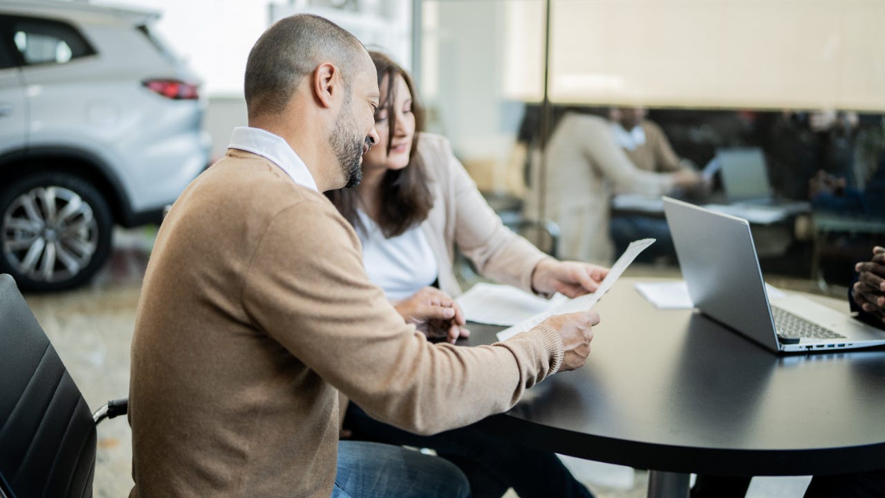 Man and woman sitting in dealership reviewing papers at a table across from salesperson who is out of frame