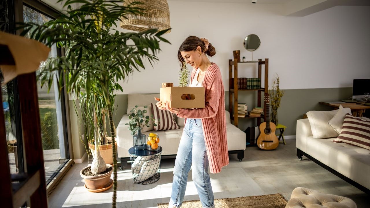 Young white woman carrying a box while walking around an open living room space.