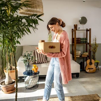 Young white woman carrying a box while walking around an open living room space.