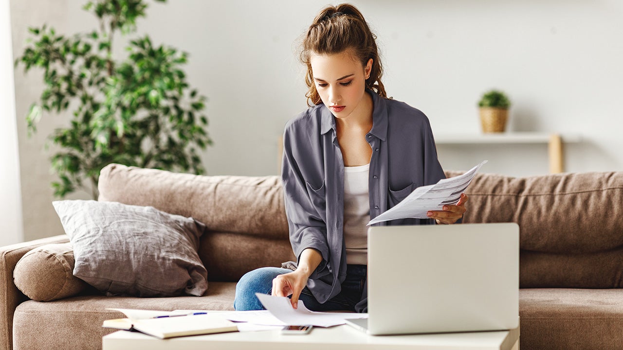 Calm focused female freelancer in casual clothes revising reports while sitting on soft couch at table and using computer in light contemporary apartment