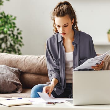 Calm focused female freelancer in casual clothes revising reports while sitting on soft couch at table and using computer in light contemporary apartment