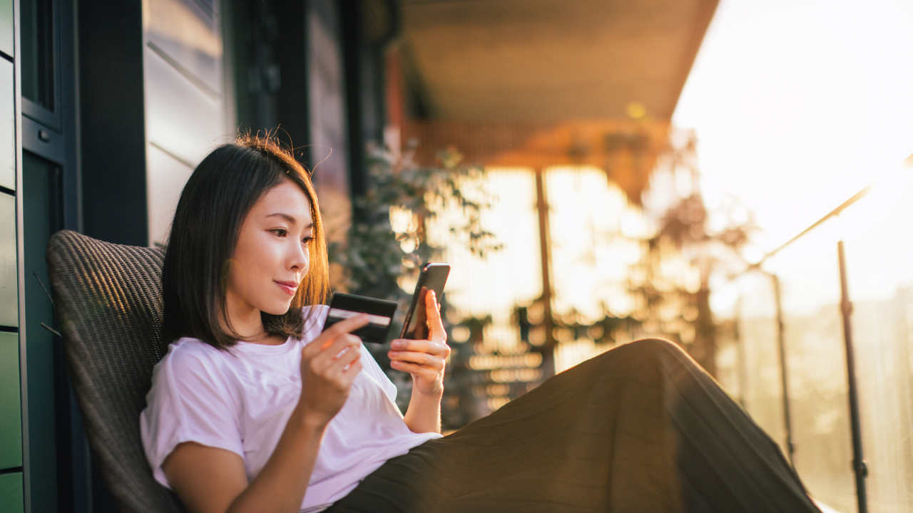 Smiling young Asian woman using smart phone and credit card to mange online banking, sitting on the balcony with dramatic sunlight. Online shopping makes life easier.