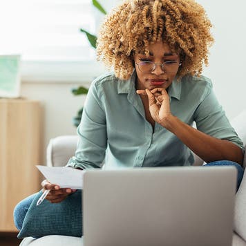 Serious woman is sitting on a sofa and looking at a laptop while holding a paper in her hand. She is wearing glasses. She might be working from home or studying, or paying a utility bill. She is wearing a green shirt and jeans.