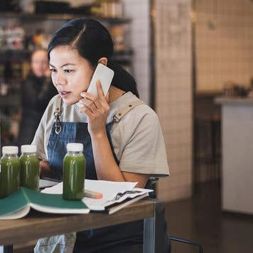 Asian female entrepreneur talking over smart phone while working on laptop in store