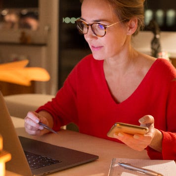 Woman wearing glasses on laptop while holding phone