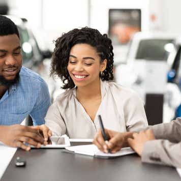 Spouses Signing Papers With Auto Seller In Dealership Office